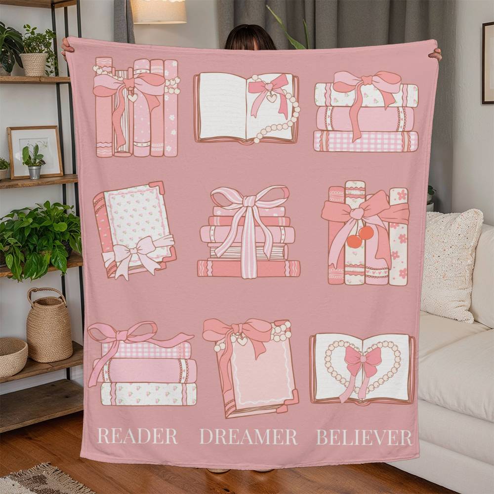 Pink fleece blanket with coquette-style grid of books and bows, and the words 'Reader, Dreamer, Believer' at the bottom.
