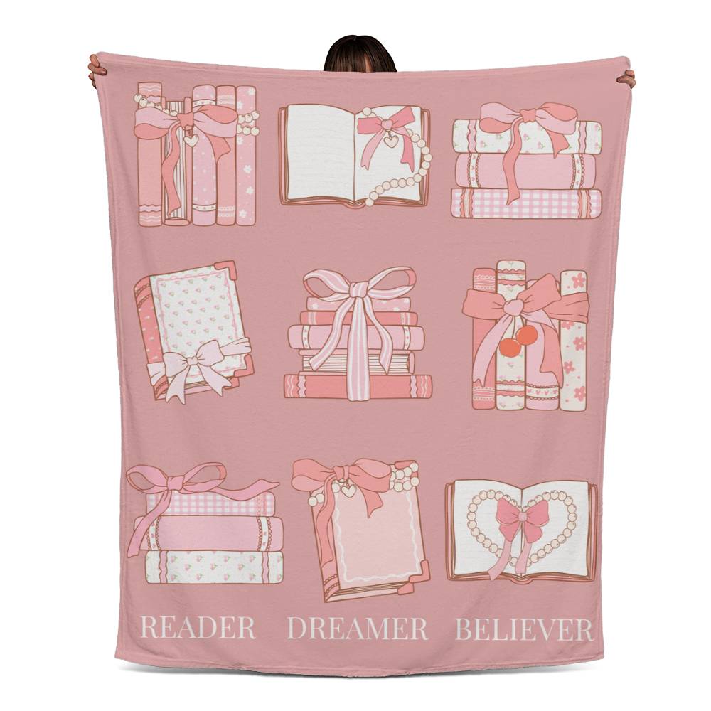 Pink fleece blanket with coquette-style grid of books and bows, and the words 'Reader, Dreamer, Believer' at the bottom.
